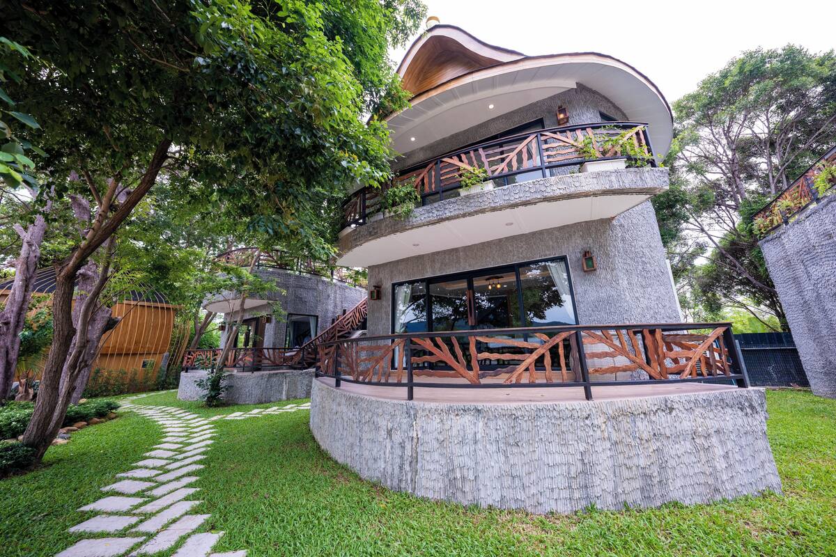A modern two-story house with a curved roof, a stone facade, and a balcony with wooden railings.