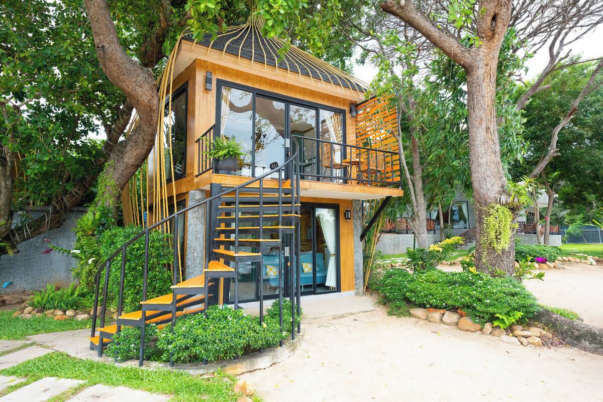 A treehouse with a wooden exterior, glass windows, and a staircase leading to a balcony.