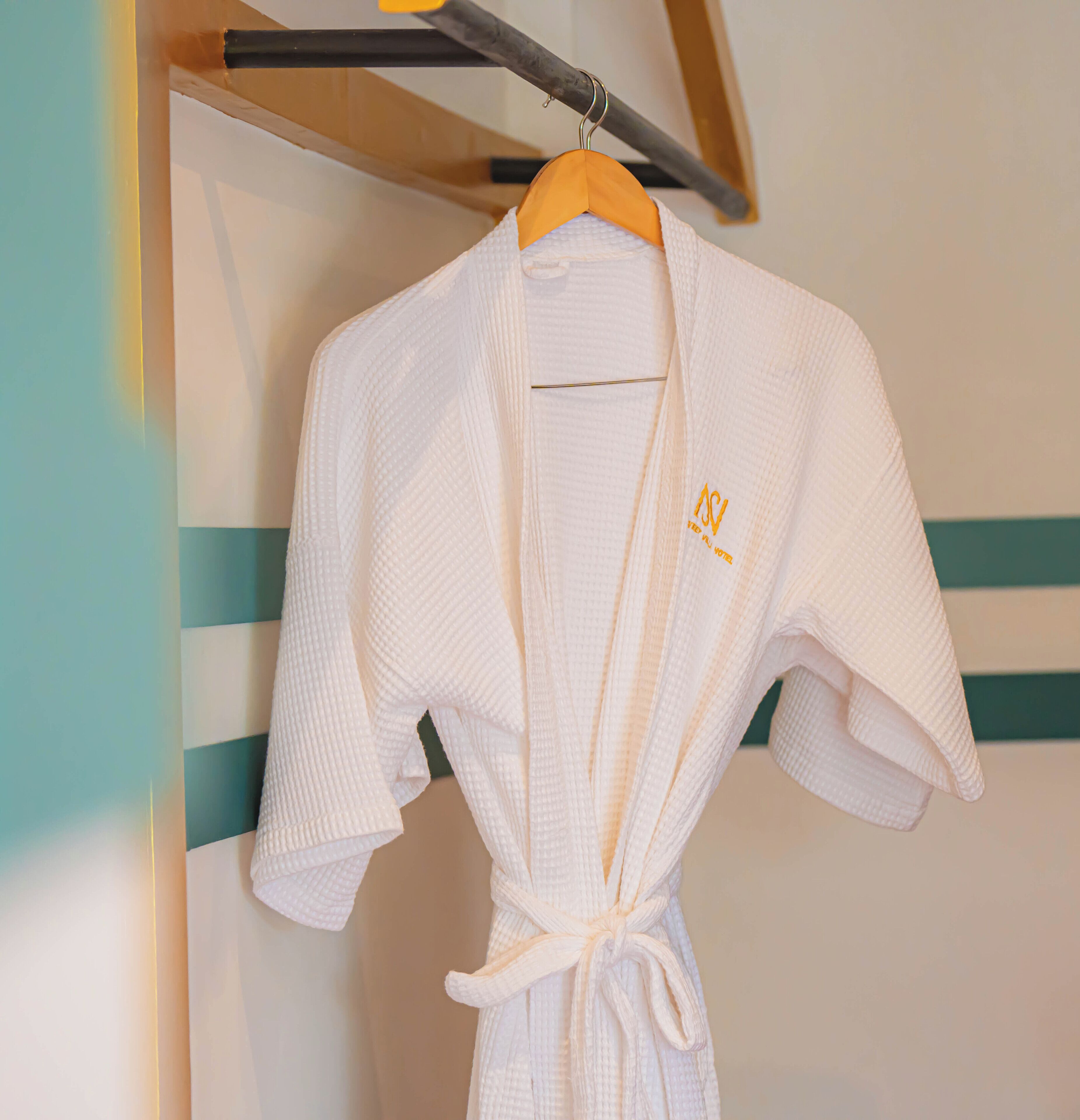 A white robe with a tied knot at the waist, hanging on a wooden hanger against a wall with a striped pattern.