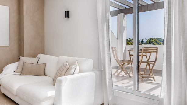 A white sofa with striped pillows, a wooden table with chairs, and a view of a coastal landscape through a glass door.