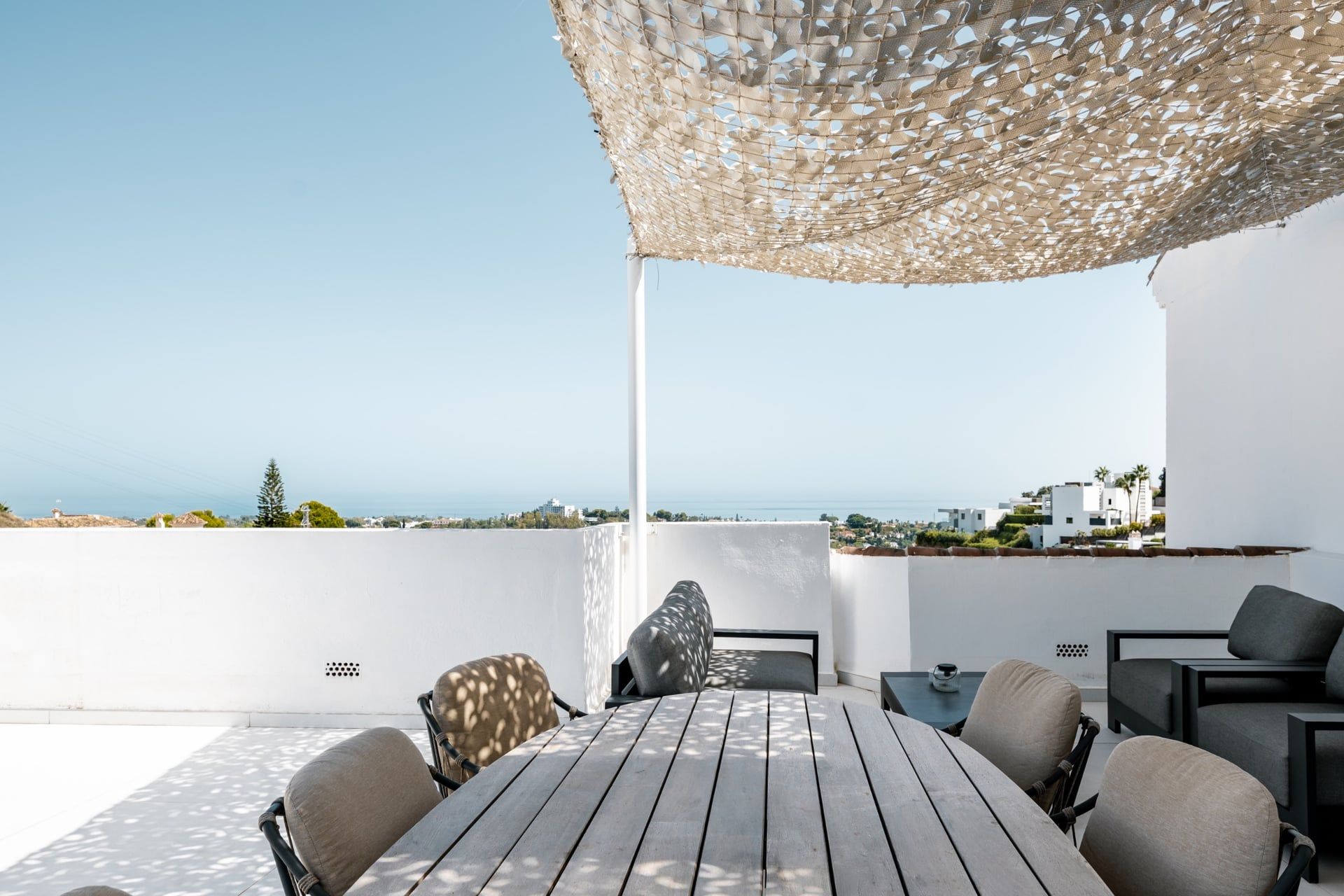 Une terrasse extérieure avec une table et des chaises en bois, un auvent en fibres tressées et une vue sur la mer et les bâtiments.
