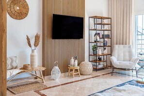 A modern living room with a flat-screen TV, a wooden slatted wall, a white armchair, a tall black and wood shelving unit, and a patterned rug.