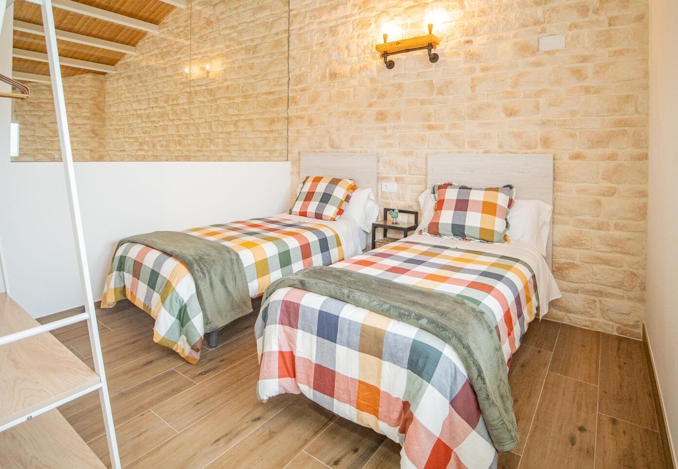 A bedroom with two beds, a wooden ceiling, stone walls, and a small nightstand with a lamp.