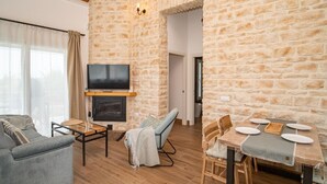 A cozy living room with a stone wall, a fireplace, a TV, a sofa, a dining table with chairs, and a window with curtains.