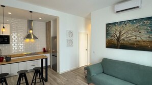 A modern kitchen with white tiles, a stainless steel range hood, and pendant lighting. There is a dining area with a green sofa and a coffee table. A painting of a tree is on the wall.