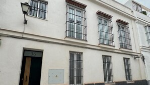 A building with a white facade, black lower section, and barred windows. There is a door labeled '36' and two electrical boxes with warning symbols.