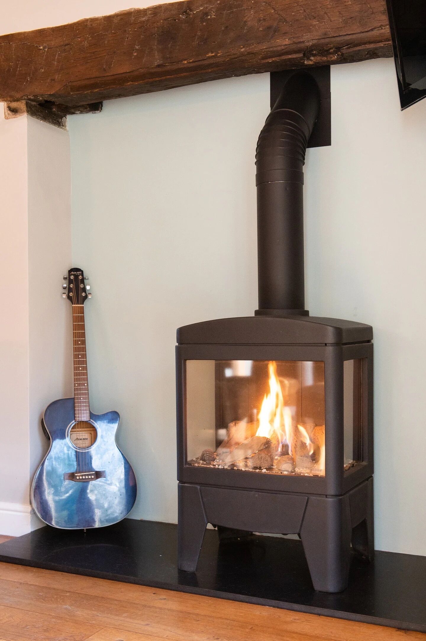 A black wood-burning stove with a glass front, a guitar, and a wooden beam above it.