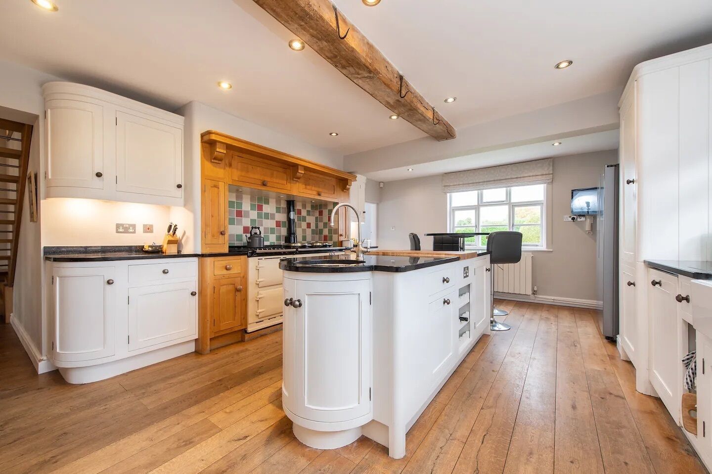 A spacious kitchen with white cabinetry, a central island, wooden beams on the ceiling, and a large window providing natural light.
