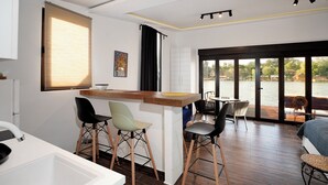 A modern kitchen with a bar area, two bar stools, a dining table with chairs, and a large sliding glass door leading to a balcony with a view of the water. - Beach House Belgrade (Belgrade)