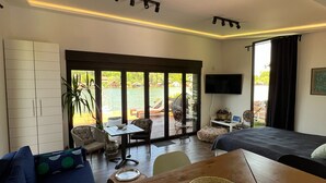 A modern living room with a sofa, a dining table, and a TV. There is a large window with a view of the outdoors. - Beach House Belgrade (Belgrade)