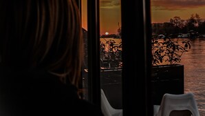 A person seated by a window overlooking a sunset over a body of water with boats. - Beach House Belgrade (Belgrade)