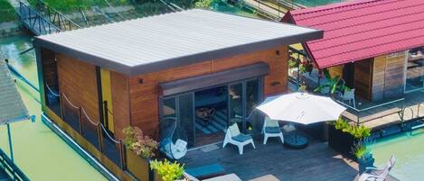 Una casa flotante moderna con un porche cubierto, mobiliario exterior y un muelle que se adentra en una masa de agua verde.
