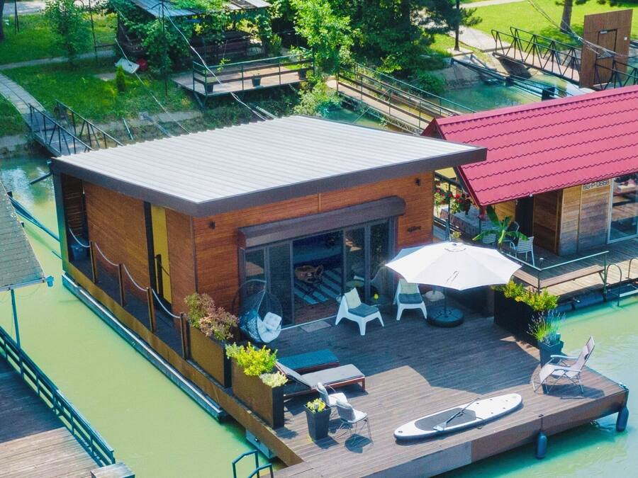 A modern houseboat with a covered patio, outdoor seating, and a dock extending into a green body of water.