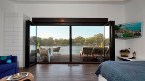 A bedroom with a large sliding glass door leading to a balcony with a view of a lake and outdoor furniture. - Beach House Belgrade (Belgrade)
