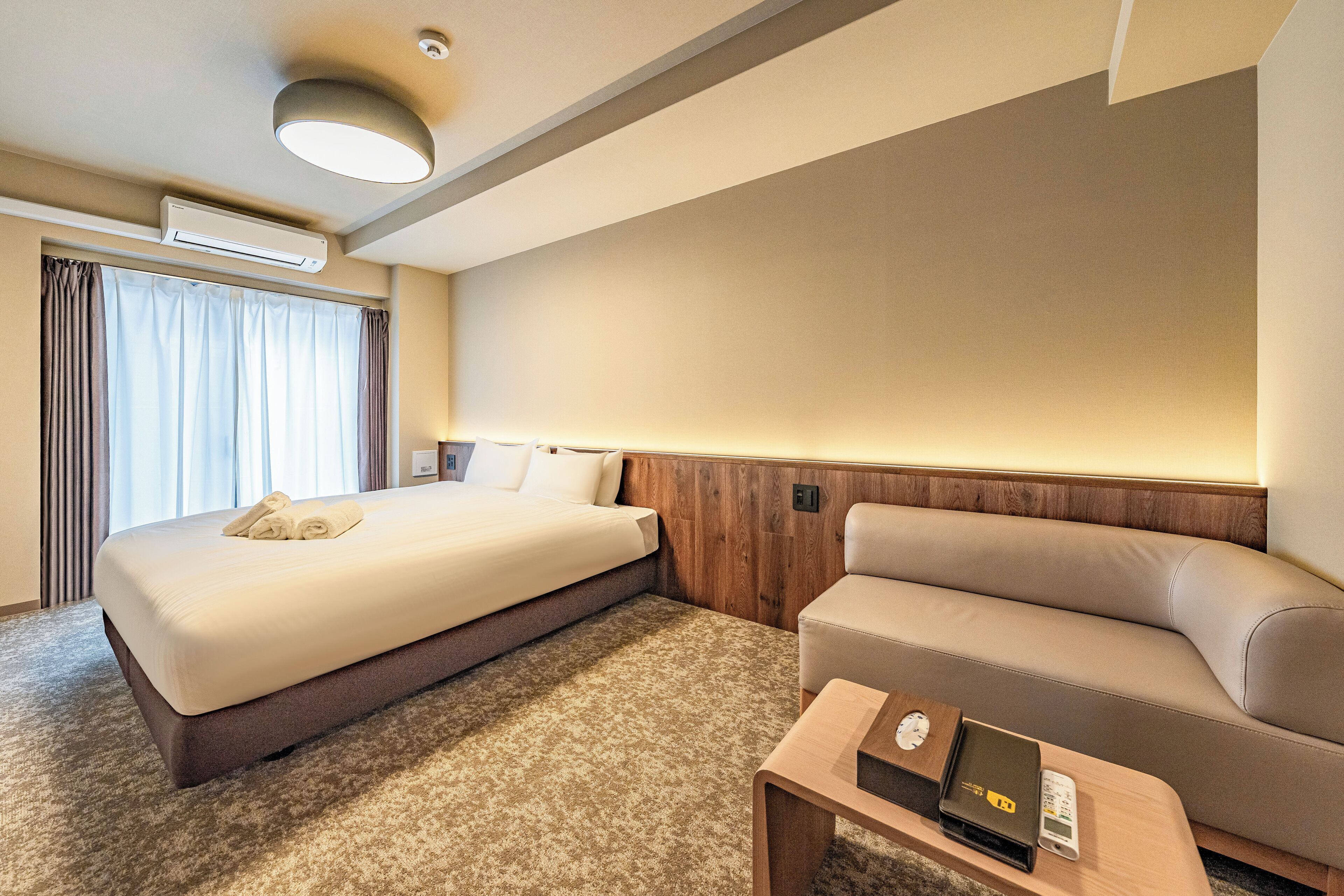 A hotel room with a large bed, a sofa, a small table with a clock, and an air conditioning unit.