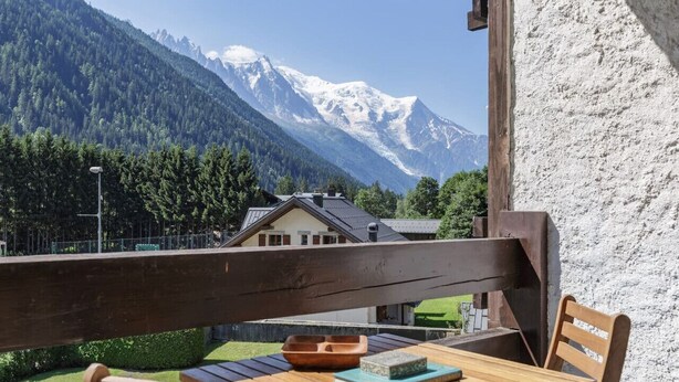 Una terraza de madera con una mesa y sillas, con vistas a un paisaje montañoso con picos nevados y una casa al fondo.