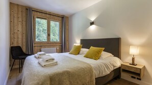 Un dormitorio con una cama grande, dos almohadas, una mesita de noche con una lámpara, una silla y una ventana con cortinas.
