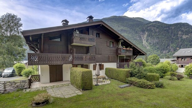 A two-story wooden house with a balcony, surrounded by well-maintained hedges and a grassy lawn.