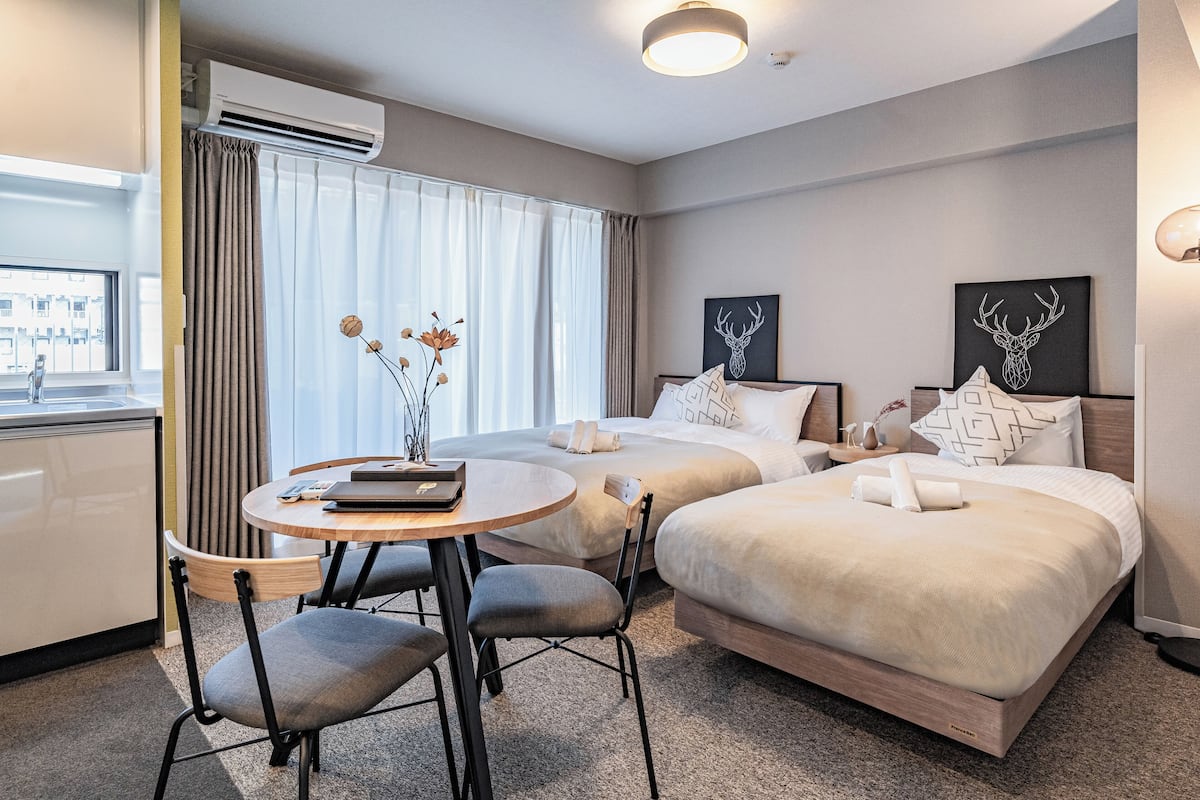 Quarto de hotel com duas camas, uma mesa de jantar com cadeiras, ar condicionado e uma janela com cortinas.