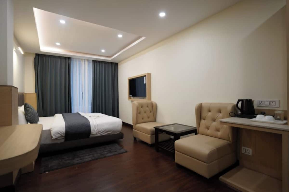 A modern hotel room with a bed, two armchairs, a small table, a TV, and a coffee station.