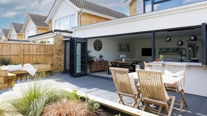Une terrasse extérieure moderne, aménagée avec du mobilier en bois, une table à manger et un coin cuisine équipé d’un évier et d’un plan de travail.