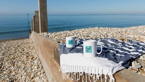 Un banc en bois, deux tasses blanches sur lesquelles est inscrit « Air frais » et une serviette bleue et blanche à rayures, le tout posé sur une plage de galets.