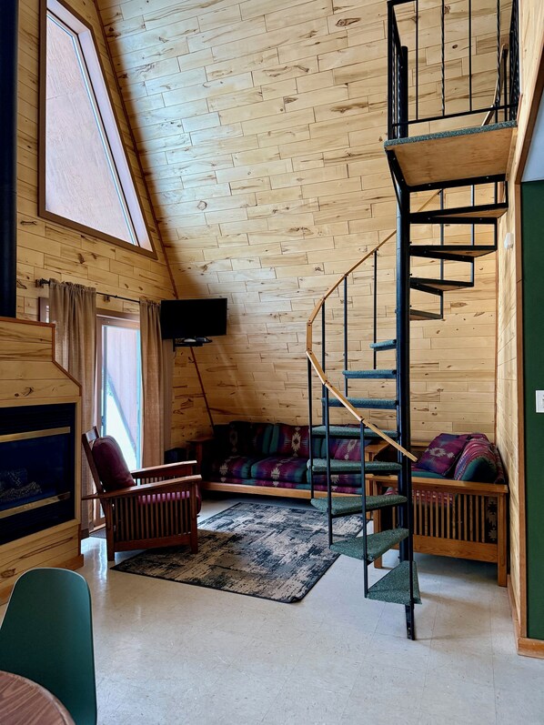 Une pièce de style « loft » avec un plafond en pente en bois, une cheminée intégrée, un canapé vert, un escalier et un téléviseur à écran plat fixé au mur.