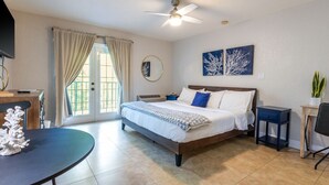 Ein Schlafzimmer mit Bett, Deckenventilator, Fenster mit Vorhängen, kleinem Tisch und einer Pflanze.