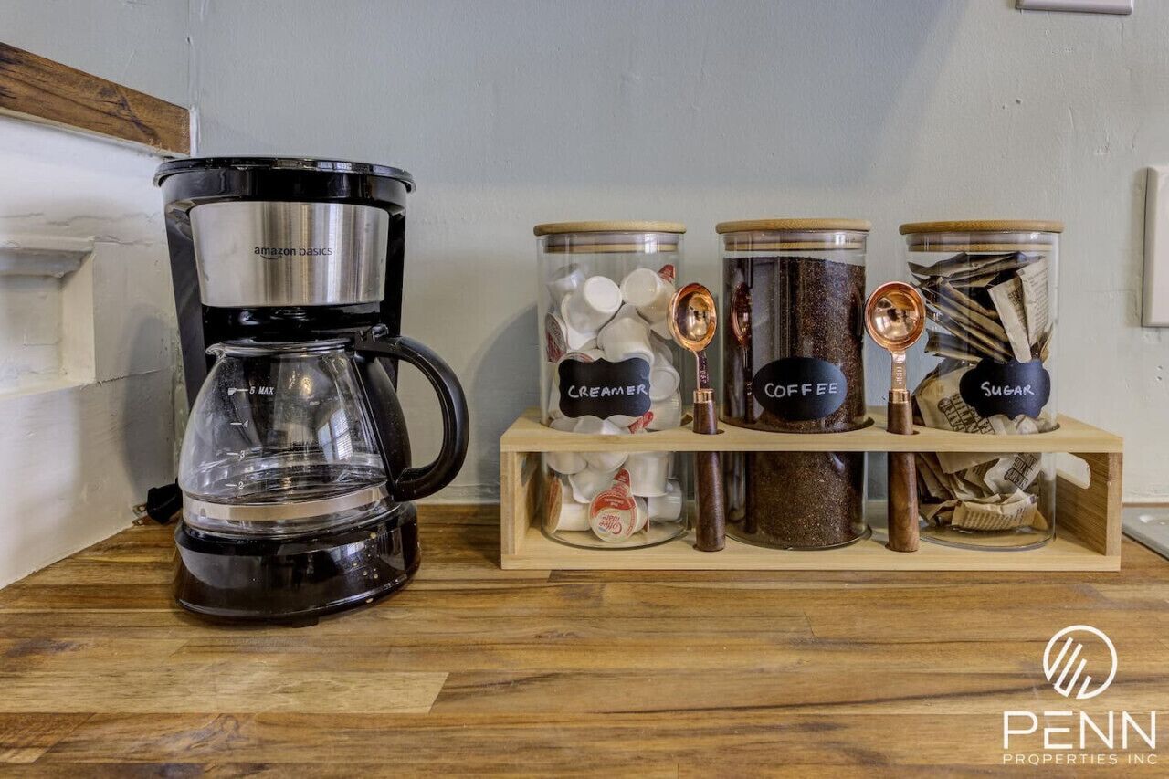 Uma cafeteira, três recipientes identificados com grãos de café, açúcar e leite, e uma prateleira de madeira.