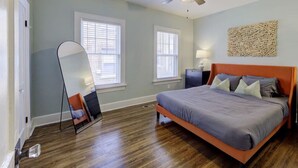 Un dormitorio con una cama, ventilador de techo, dos ventanas, una mesita de noche y un espejo montado en la pared.