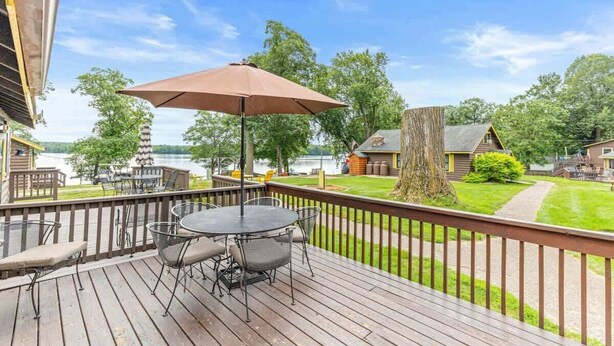 Une terrasse en bois avec une table et des chaises, un grand parasol, et une vue sur un lac et des b âtiments en arrière-plan.