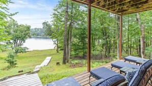 A covered outdoor seating area with wooden deck and lounge chairs, overlooking a lake and forested area. - Black Squirrel Retreat (Minong)