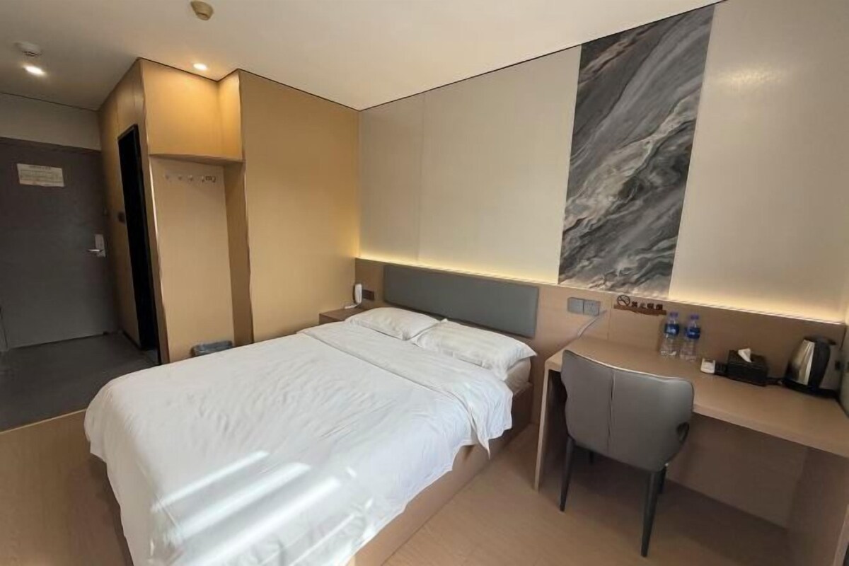 Quarto de hotel com cama, escrivaninha, cadeira, mesa de cabeceira e uma obra de arte na parede.