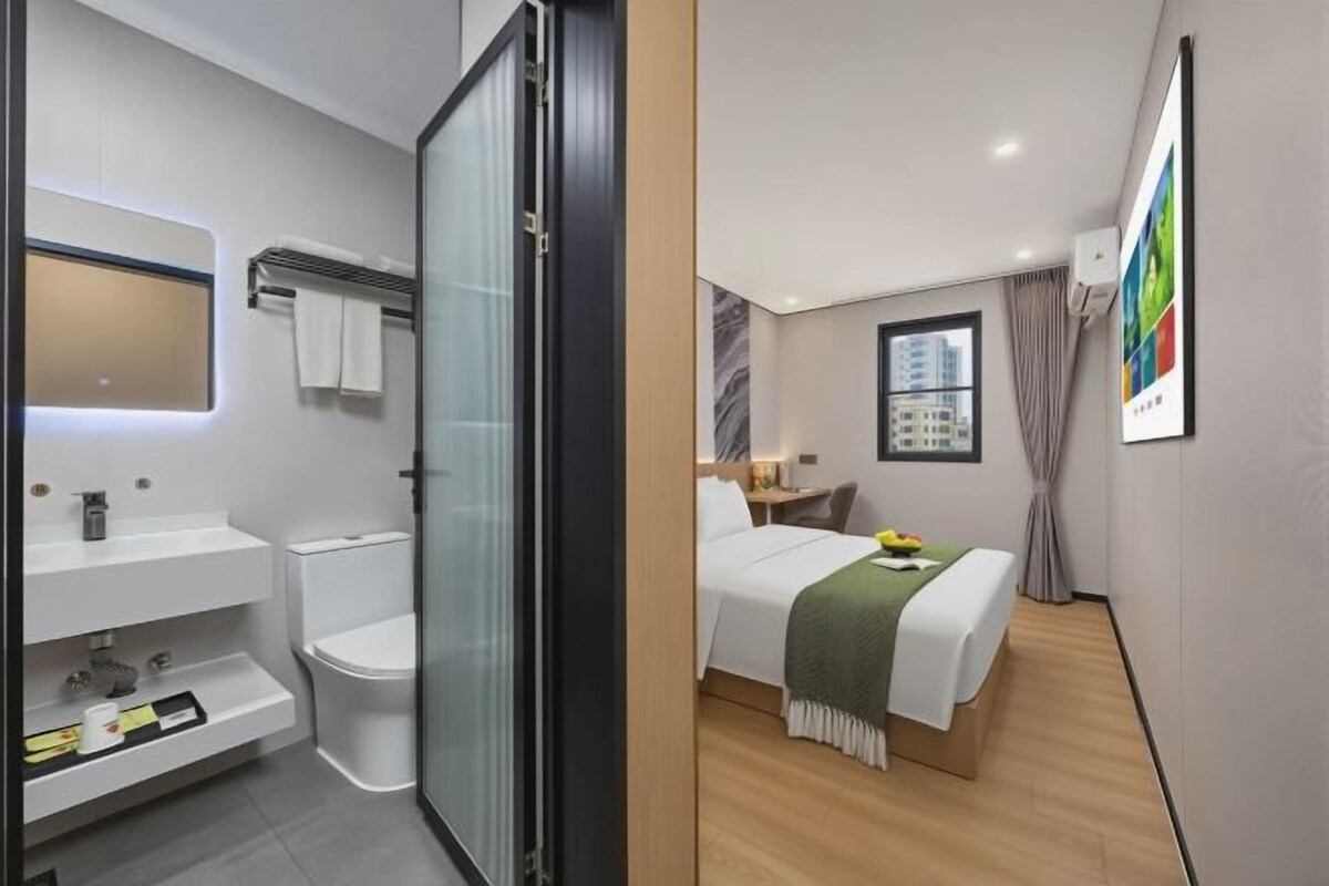 A modern hotel room with a bathroom, a bed, a bedside table, and a window.
