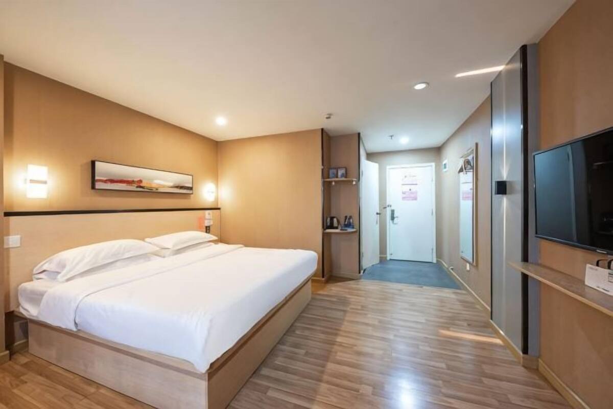 A hotel room with a bed, a television, a door, and a hallway.