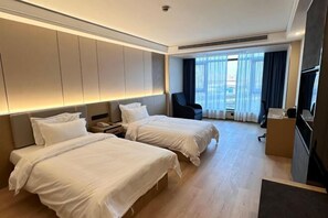 Habitación de hotel con dos camas, un escritorio, una silla, un televisor y un ventanal con cortinas.