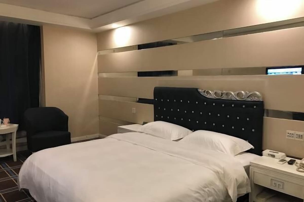 A hotel room with a bed, bedside table, chair, and a wall-mounted control panel.