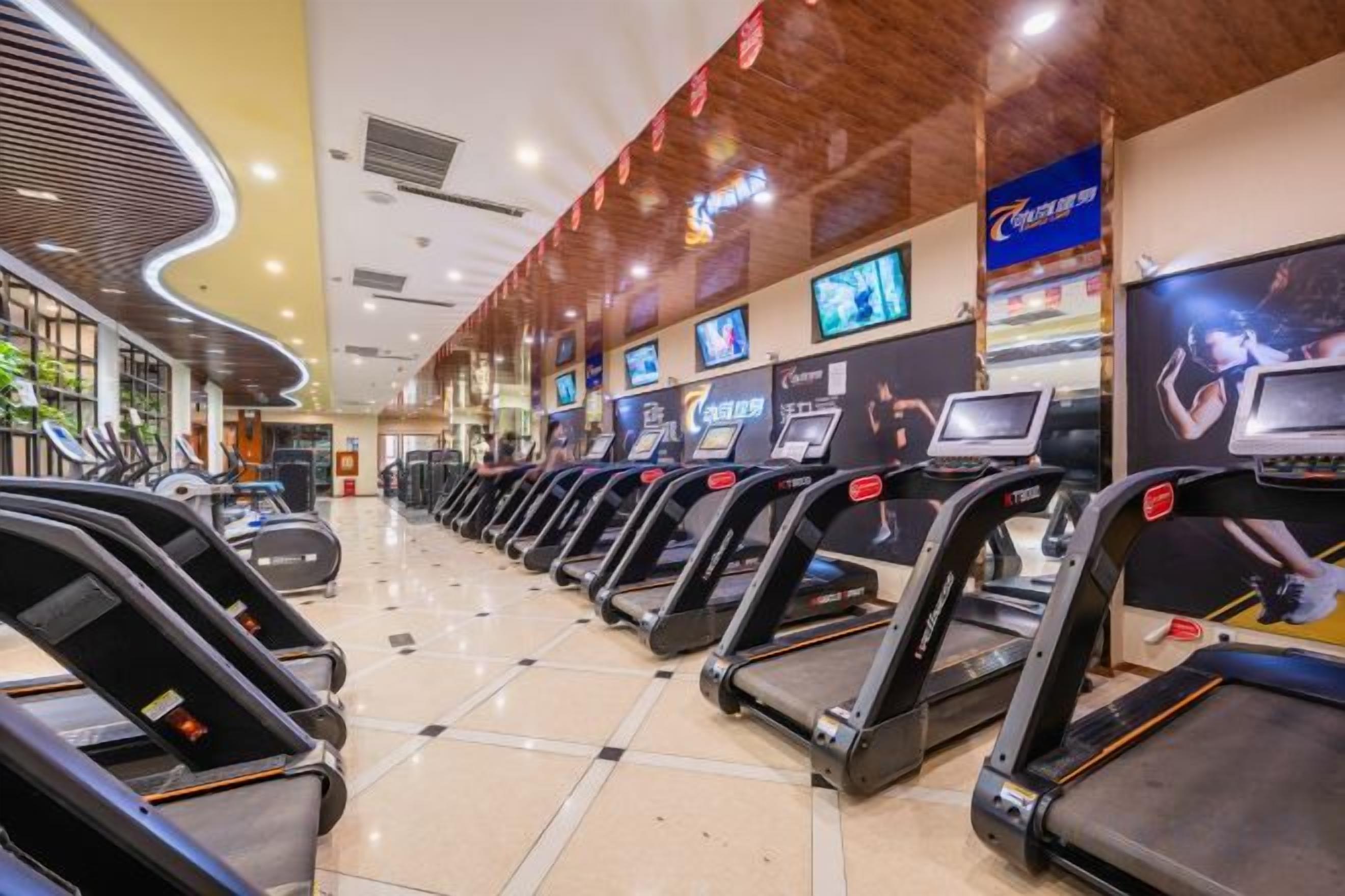 Une salle de sport avec des tapis de course alignés en rangées, un plafond en bois et un grand écran diffusant une publicité.