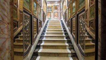 Un magnifique escalier en marbre, agrémenté de décorations élaborées, mène à un espace aux hauts plafonds où sont exposées des œuvres d’art encadrées.