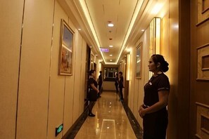 A hotel corridor with a polished floor, framed artwork on the walls, and a uniformed staff member standing.