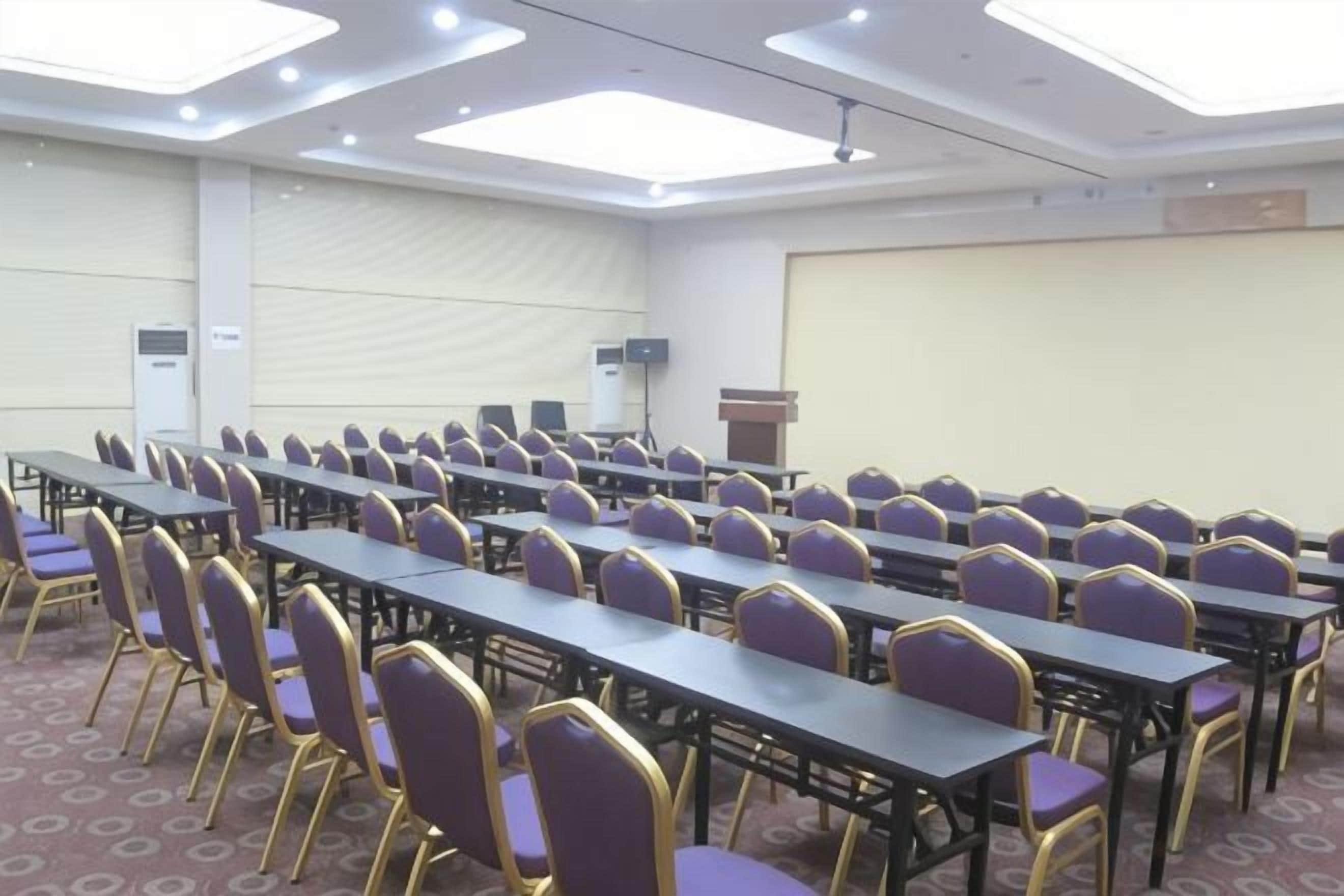 Une salle de conférence avec des rangées de tables et de chaises, un pupitre et un écran de projection.