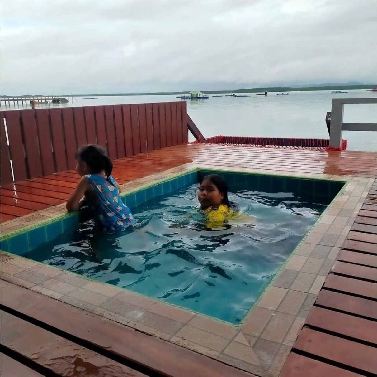A swimming pool with two children playing, surrounded by a wooden deck and a view of the sea.