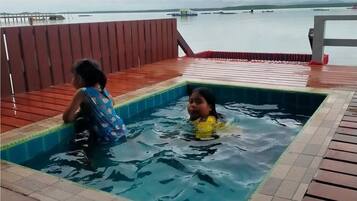 A swimming pool with two children playing, surrounded by a wooden deck and a view of the sea.