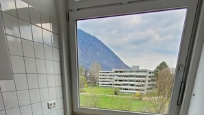 A kitchen with a built-in stove, white cabinets, and a window overlooking a green landscape and a building. - Apartment 304, apartment, 38 sqm, 1-2 persons, balcony (Bad Reichenhall)