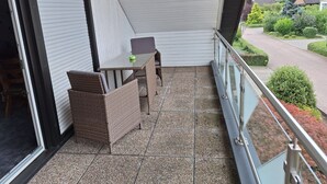 Ein Balkon mit Tisch, Stühlen und einer Korbsitzbank, mit Blick auf eine Wohnstraße.