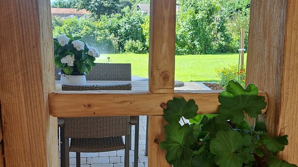 Une fenêtre à cadre en bois donnant sur un jardin, une table avec des chaises et une plante en pot.