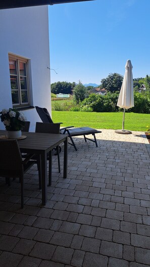 Een terras buiten met een tafel en stoelen, een overdekte parasol en uitzicht op een groen landschap.