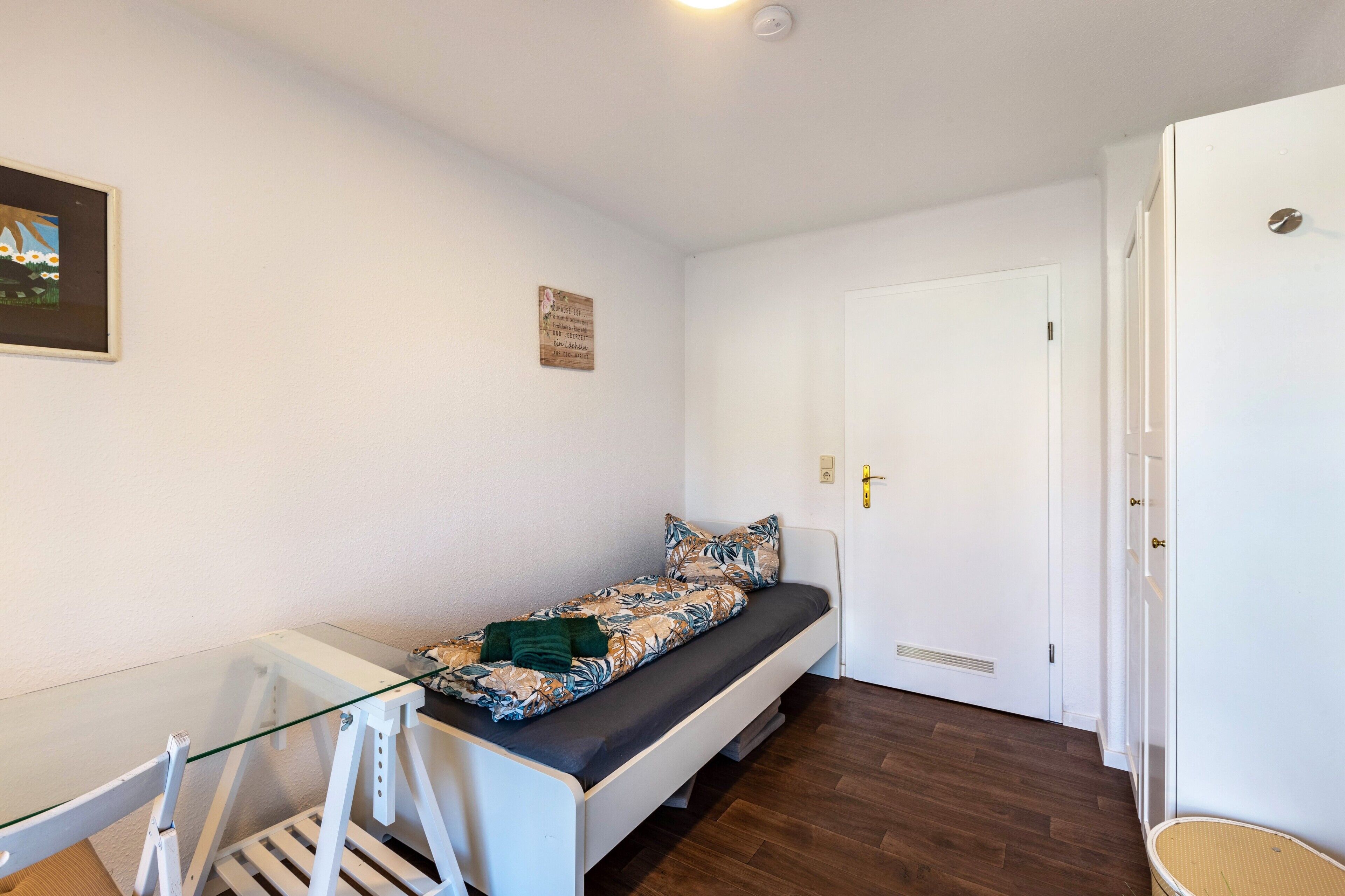 Ein kleines Zimmer mit einem Einzelbett, einem Glastisch, einem weißen Stuhl, einer Tür und einem an der Wand hängenden Bild.