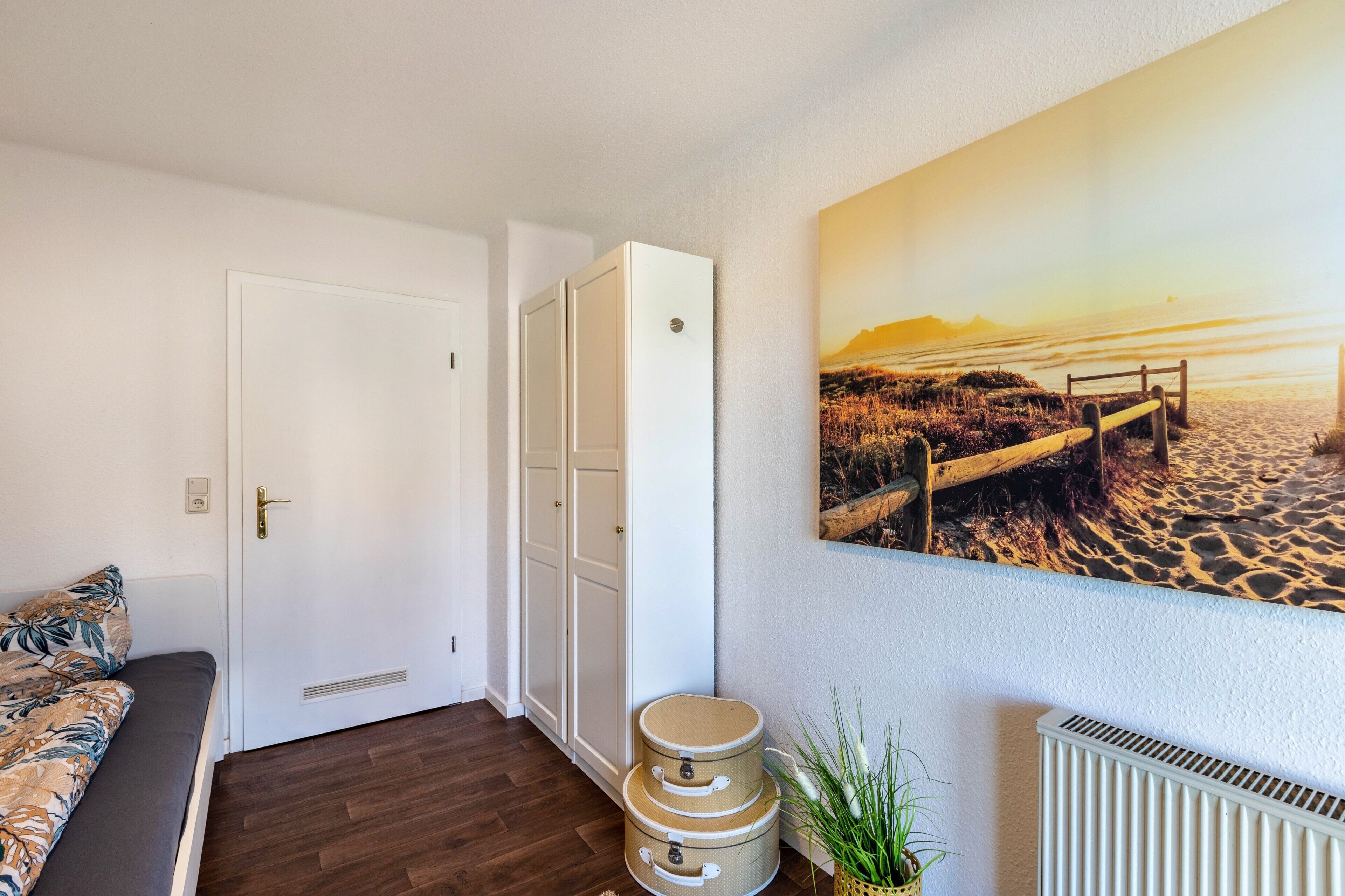 A room with a bed, a white door, a white wardrobe, a painting of a beach, and a radiator.