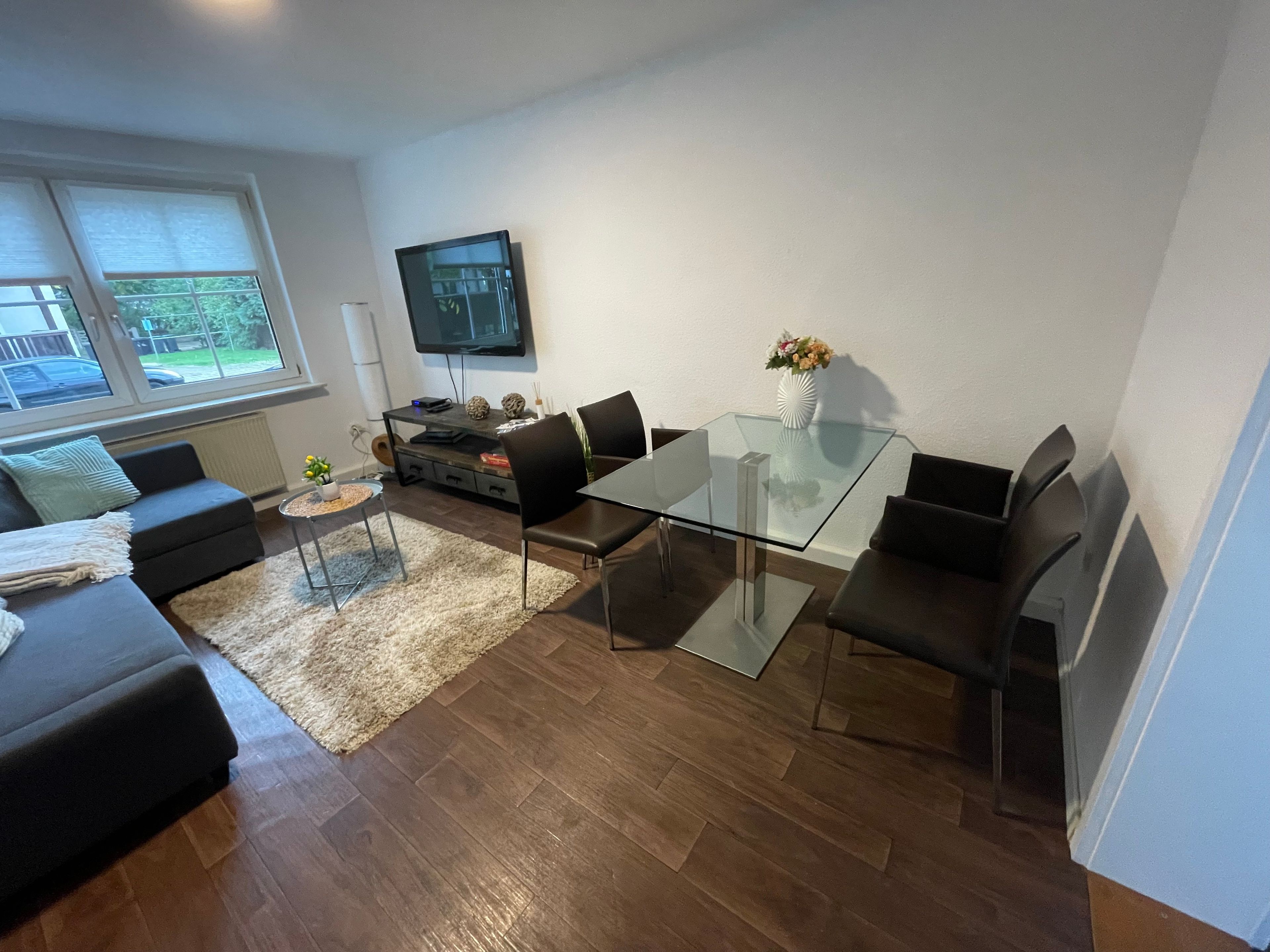 Una sala de estar moderna con una mesa de comedor de vidrio, sofás negros, un televisor montado en la pared y vistas al exterior a través de amplios ventanales.